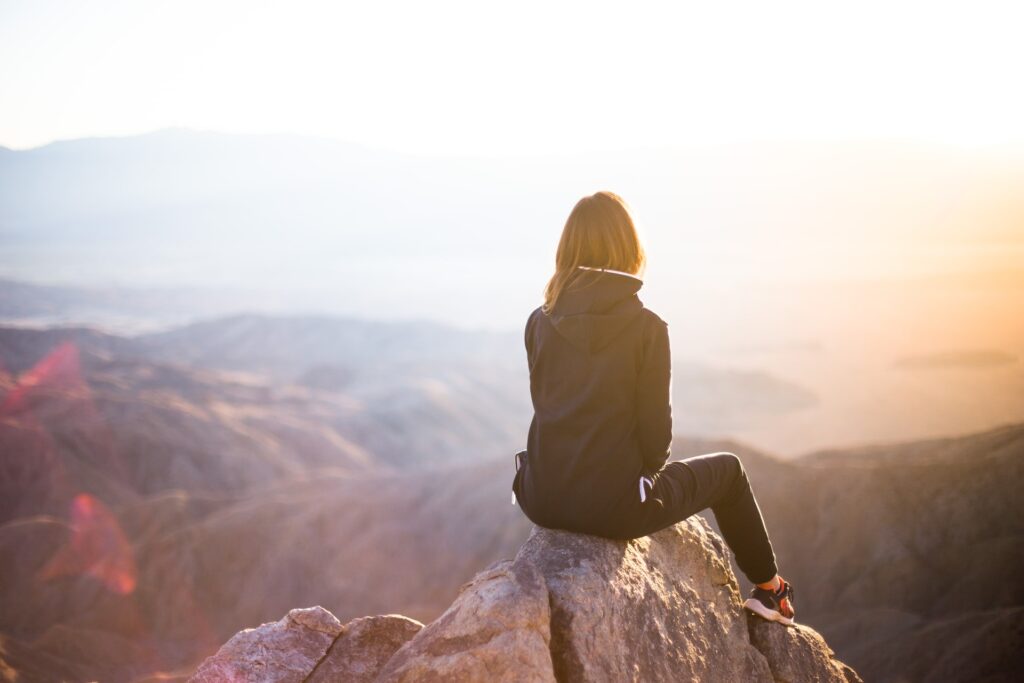 A woman sitting on top of a rocky outcrop with her back to the camera and sun streaming across the landscape